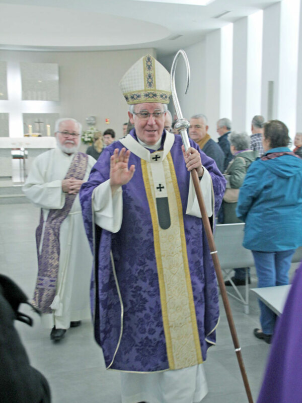 Imagen de un sacerdote en vestimenta religiosa, posiblemente durante una ceremonia o celebración. El sacerdote lleva un manto azul con detalles dorados y una mitra blanca. La imagen muestra a varios otros individuos en el fondo, algunos también vestidos con ropa religiosa. El ambiente parece ser interior, posiblemente en una iglesia o edificio dedicado a la religión.