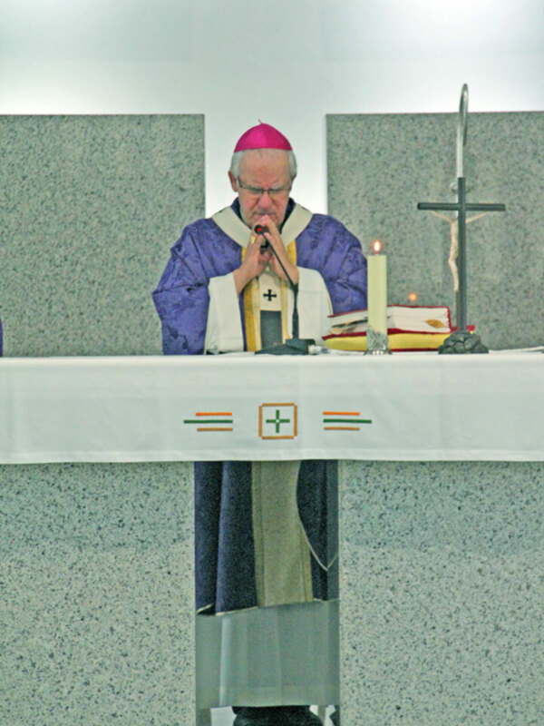 Un sacerdote vestido con un hábito de color azul y blanco, lleva una cruz en el pecho. Está frente a un altar con una vela encendida y un crucifijo en el fondo. La imagen muestra a la persona orando, con una expresión serena y profunda en su rostro.
