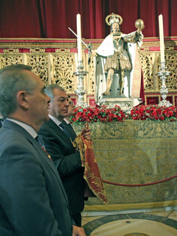 Dos hombres en traje se encuentran frente a una estatua de un rey, rodeados por flores rojas y velas encendidas. La escena se desarrolla en un entorno decorado con detalles artísticos y una alfombra circular.
