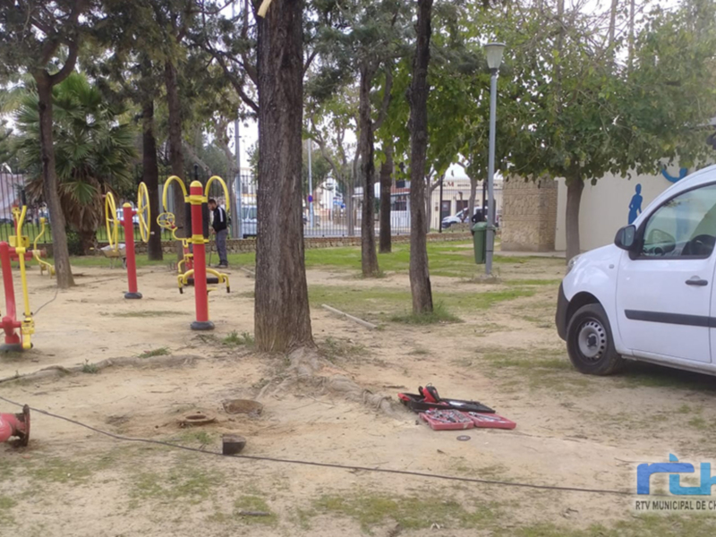 Vehículo blanco estacionado cerca de un parque infantil con juegos de color rojo y amarillo, y un árbol en el centro. El parque está rodeado por una cerca metálica y hay un poste de luz cerca del árbol.