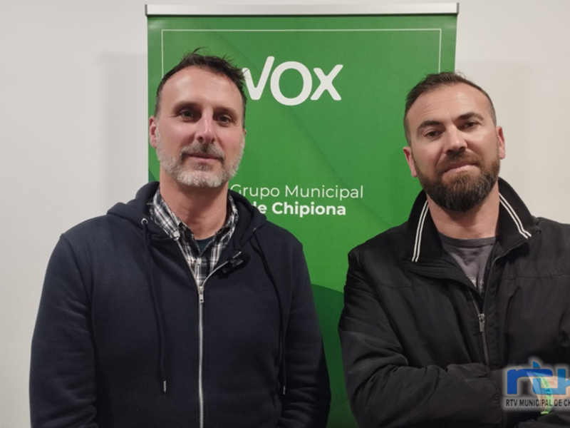 Dos personas posando frente a una banderola verde con el logotipo de VOX y el nombre de un grupo municipal.