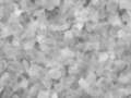 Una vista microscópica de partículas o cristales en blanco y negro, posiblemente una muestra de material mineral. La textura es irregular con formas que parecen hojas o láminas, lo que sugiere una estructura compleja en el material.