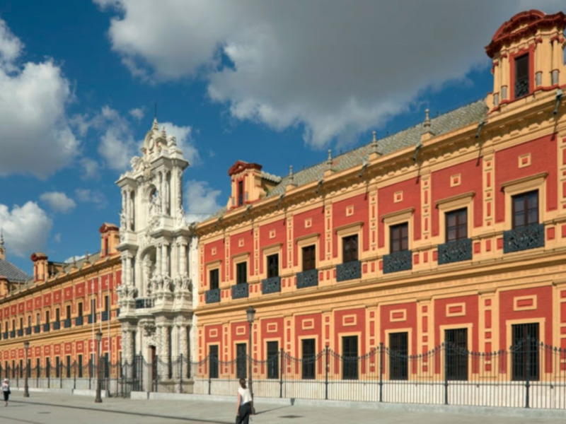Imaginación: Un edificio histórico con una torre central y ventanas cuadradas, rodeado por un muro de hierro forjado bajo un cielo azul con nubes. El edificio parece ser de estilo neoclásico o barroco, con detalles ornamentales en la parte superior.