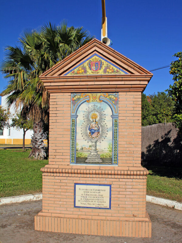 Un monumento religioso con una imagen de la Virgen María y un escudo heráldico, rodeado de azulejos y un pedestal de piedra, en un entorno verde con palmeras y un cielo azul claro.