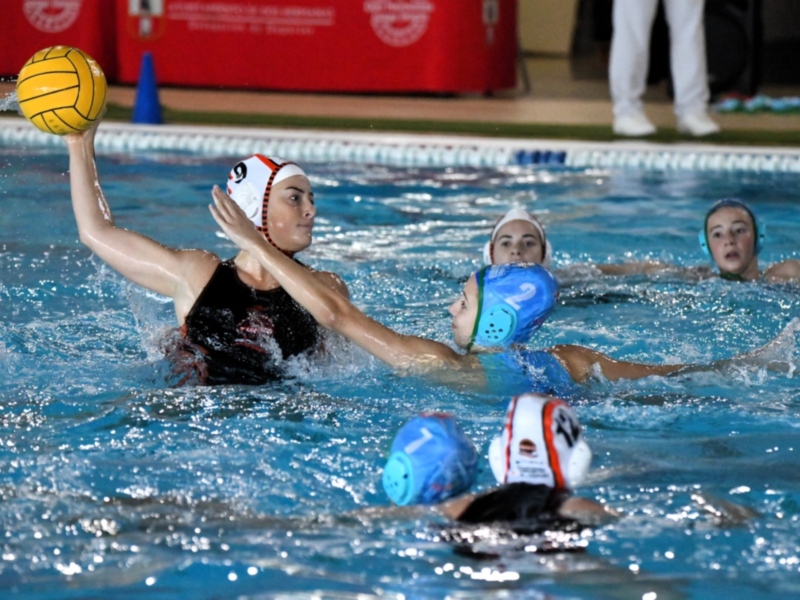 En un emocionante partido de waterpolo, las jugadoras compiten en un estadio lleno. La acción se centra en una jugadora con un casco azul que intenta derribar a otra con un casco blanco. La piscina está llena de agua azul brillante, y en el fondo se pueden ver los cascos de otros jugadores esperando su turno. La escena captura el esfuerzo y la competencia en un deporte acuático.