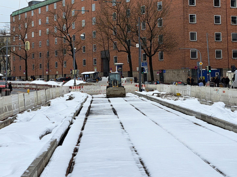 Fotografía de una calle con tránsito lento, cubierta de nieve. Vehículos y peatones circulan por una avenida con edificios de fondo. La nieve cubre el asfalto y las calles, indicando una escena de invierno. El cielo está nublado y la atmósfera parece fría.