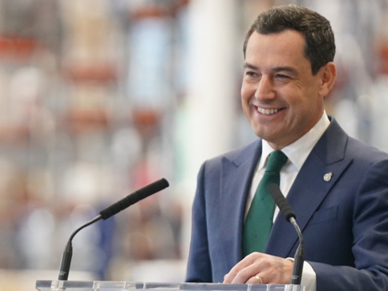 Un hombre en traje y corbata verde está hablando en un podio con un micrófono. El fondo está desenfocado, pero se pueden ver personas en el fondo.