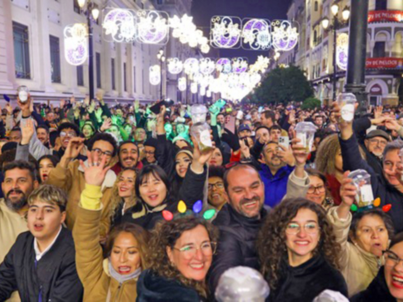 Una multitud de personas en una celebración nocturna, con luces y decoraciones en el fondo, mientras levantan sus copas al cielo.