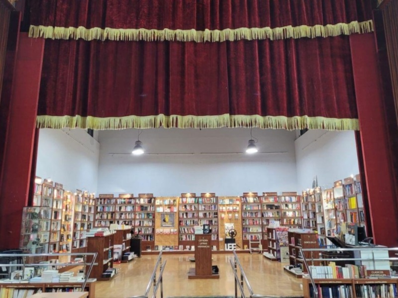 Un gran teatro con una gran cortina roja y dorada. La sala está llena de estanterías llenas de libros, iluminadas por luces blancas. La decoración y la iluminación sugieren un ambiente de lectura o conferencia.