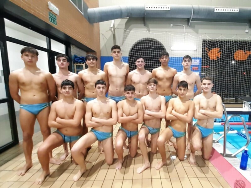 Equipo de natación masculina posando en la piscina indoor, con uniformes azules y una pared de natación en el fondo.