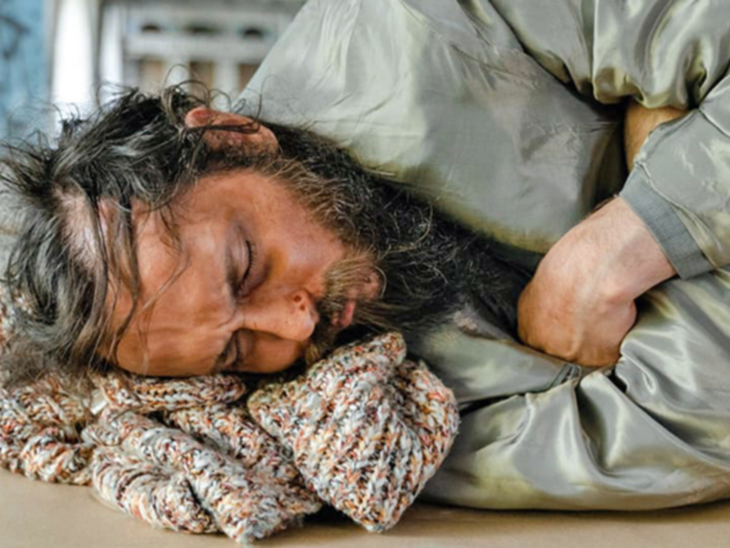 Una persona durmiendo en el suelo, cubierta con una manta, en un entorno urbano.