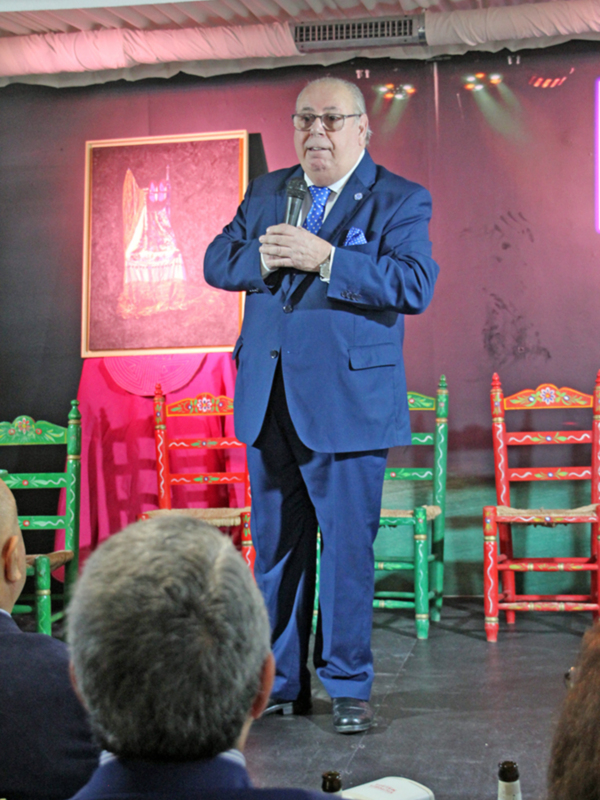 Hombre hablando en un evento formal con silla de colores en el fondo.