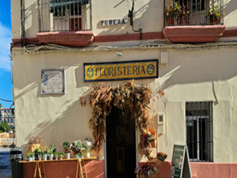 Tienda de flores en una calle de la ciudad, con un toldo y flores en el exterior. La tienda tiene una muestra de flores en la entrada. La fachada es de color blanco con detalles en rojo y azul. La calle tiene adoquines y un poste de luz en el lado.
