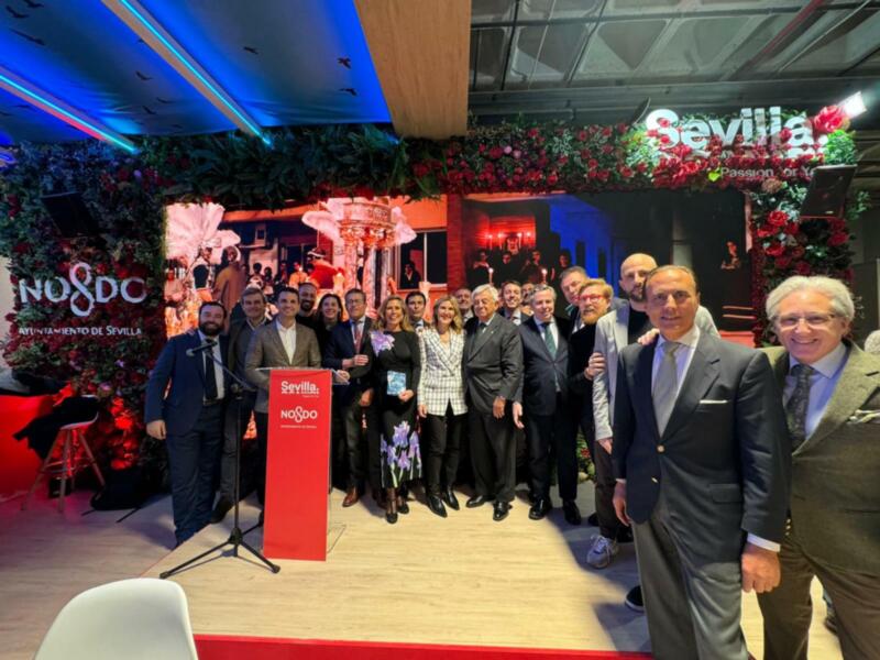 Celebración en Sevilla con un grupo de personas posando frente a una pantalla que muestra "N8DC" y "Sevilla". La imagen está ambientada en un espacio festivo con decoraciones navideñas.