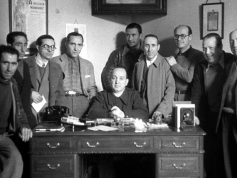 Grupo de personas en una oficina, posando para la foto. El escritorio está lleno de documentos y un teléfono. La pared detrás de ellos tiene varias imágenes y carteles colgados.