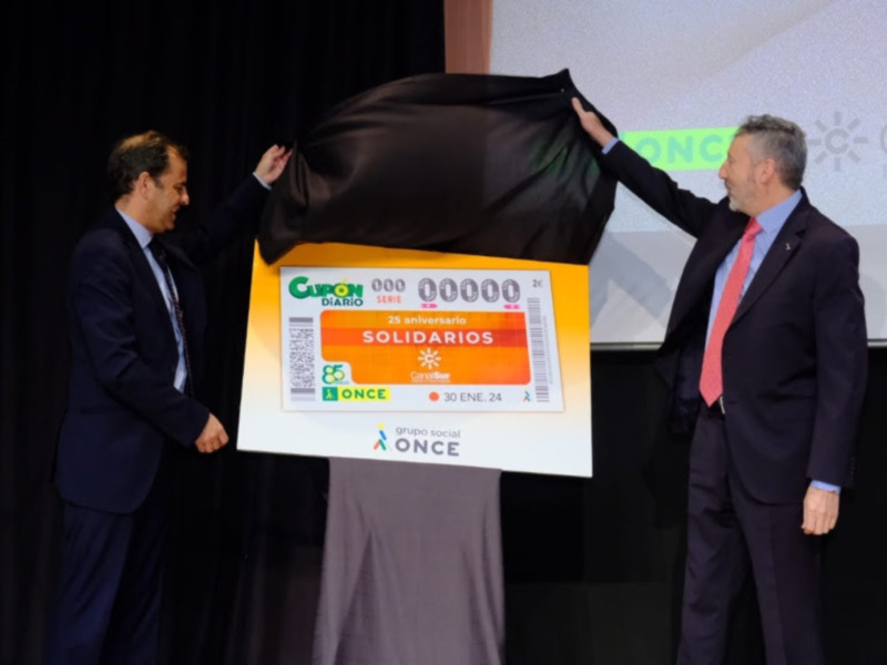 Dos hombres en trajes sostienen un gran ticket de lotería amarillo con el texto "Solidarios" y logos de ONCE. El fondo muestra un escenario con una pantalla proyectando imágenes.