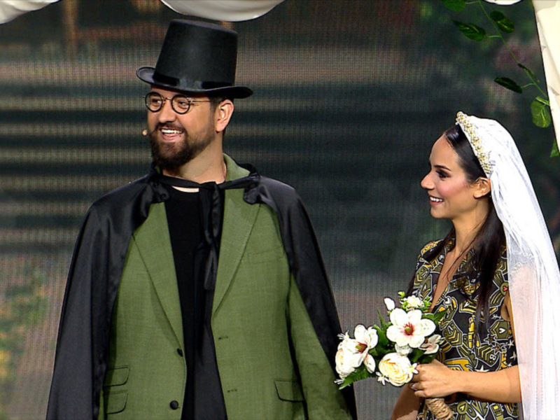 Un hombre vestido con traje verde y sombrero negro, junto a una mujer con vestido floral y velo, sonríen en una boda.