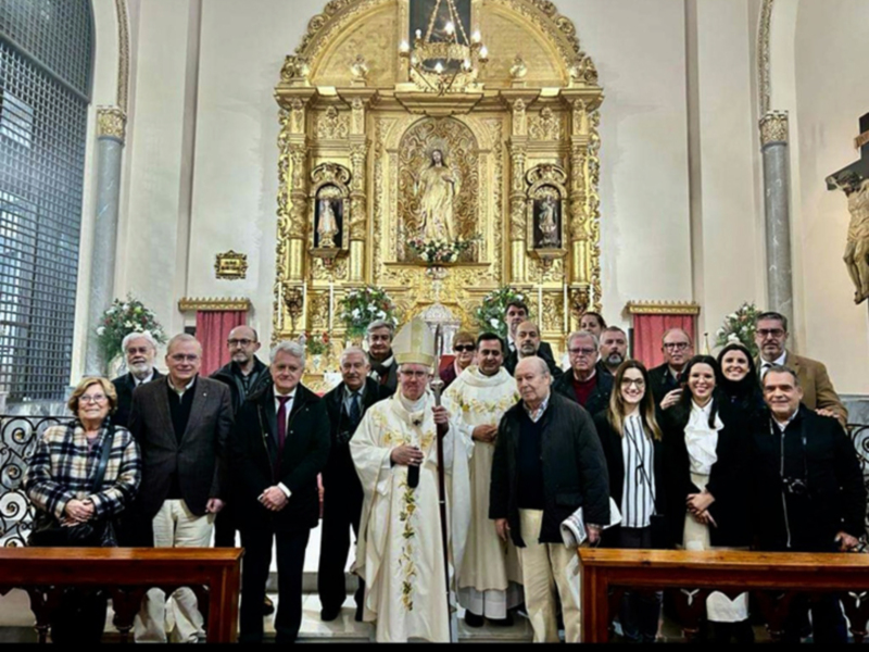 En una iglesia, un grupo de personas posa frente a un altar dorado con una imagen central. La imagen muestra a varios hombres y mujeres vestidos formalmente, incluyendo un sacerdote en traje blanco con un sombrero. La decoración dorada y la presencia de una imagen central sugieren una ceremonia religiosa o un evento oficial.