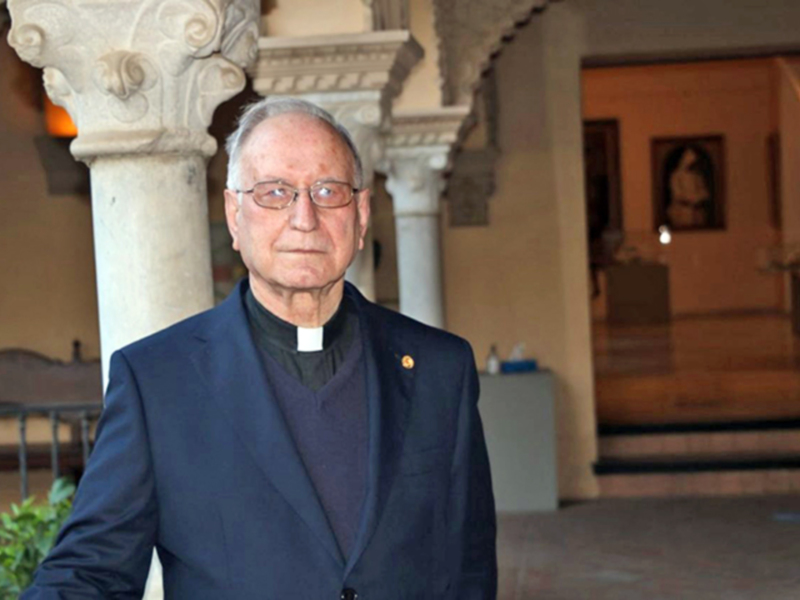 Un hombre de edad avanzada con una chaqueta azul y un cuello rojo, posando frente a una arquitectura con columnas y pinturas.