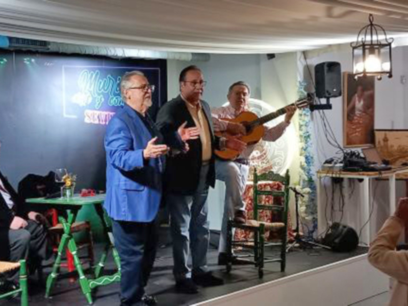 Una escena de un evento social con personas sentadas en sillas verdes y un hombre tocando guitarra en el escenario. La iluminación es cálida y la atmósfera parece ser de una reunión familiar o social.