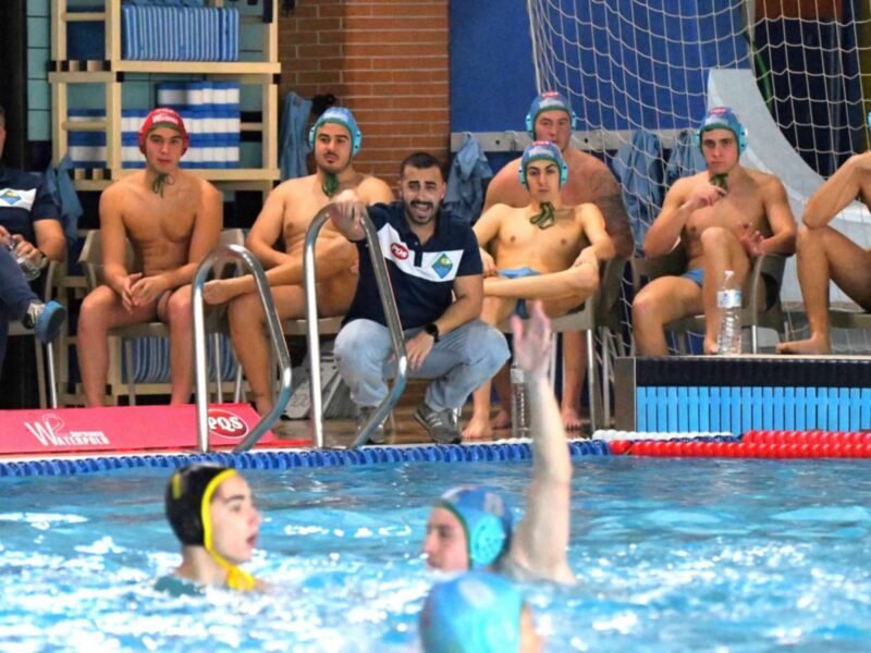 Un grupo de jugadores de waterpolo en acción en una piscina, con un árbitro y varios espectadores observando desde la orilla.