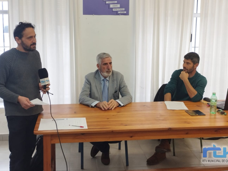 Un hombre con un micrófono en mano, otro hombre sentado en una silla y un tercero también sentado en una silla, todos frente a una mesa con documentos y un portátil.
