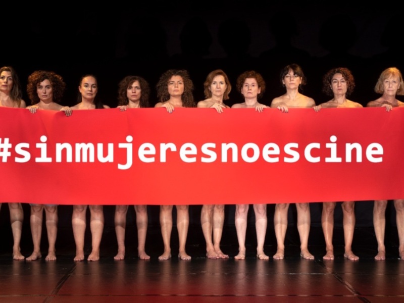 Una fila de mujeres desnudas posan en una escena teatral, detrás de ellas se encuentra un gran cartel rojo con el hashtag #sinmujeresnoescine.