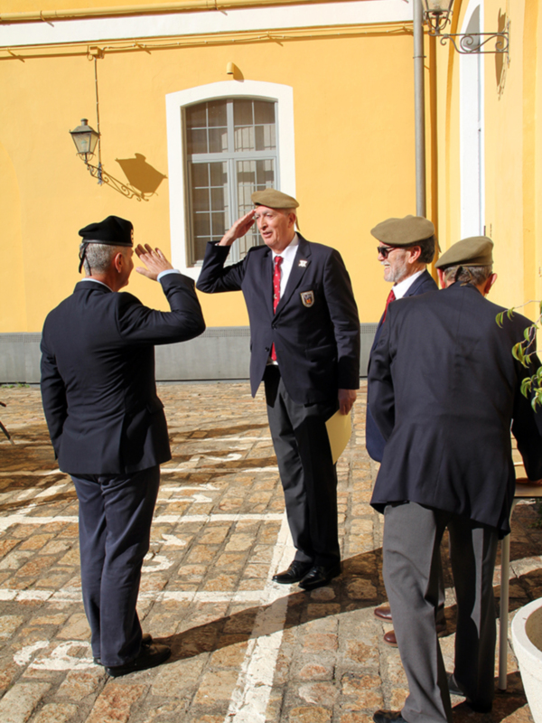 Hombres en uniforme saludando en un entorno formal con una pared amarilla y una ventana blanca en el fondo.
