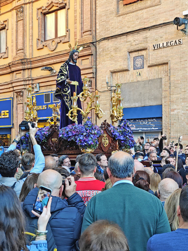 Fotografía de una procesión religiosa en una calle de Sevilla, con una imagen venerada en un carro decorado con flores y una multitud de personas observando y fotografiando el evento.