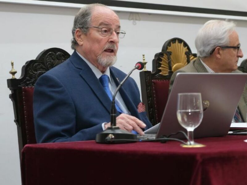 Un hombre en traje azul se dirige a una conferencia, con un portátil y un vaso de agua en la mesa. En el fondo, se puede ver una pantalla con texto en español.