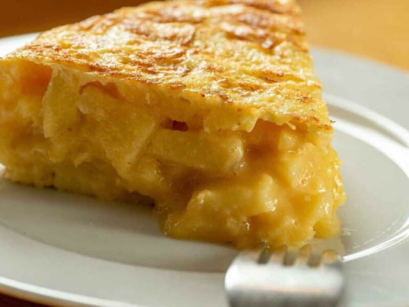 Una rebanada de tortilla española dorada y crujiente, servida en una placa blanca con un tenedor de metal. La tortilla tiene un color amarillo dorado y parece estar hecha con huevos, cebolla y otros ingredientes. La textura es crujiente en la superficie y suave por dentro, con un sabor sabroso.