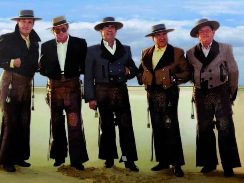 Un grupo de hombres vestidos con trajes vaqueros y sombreros de vaquero, posando en una playa arenosa con el cielo azul y nubes blancas al fondo.
