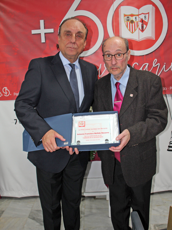 Dos hombres en trajes posan con un diploma en un evento de la Peña Sevillista San Bernardo, con un fondo rojo y blanco.