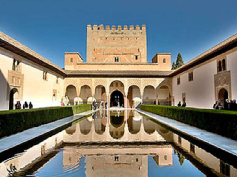 La Alhambra, un impresionante palacio fortificado en Granada, España. La vista incluye la famosa Alhambra Tower (Torre de Comares), un arco con columnas y una fuente circular. El cielo azul profundo refleja en el agua de la fuente, creando un efecto de doble imagen.