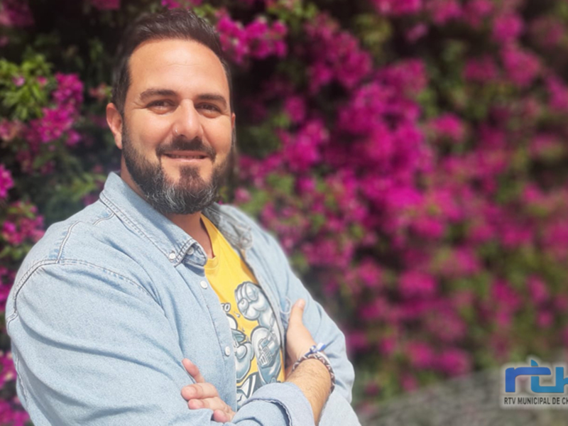 Un hombre con barba y camisa azul posando con flores en el fondo.
