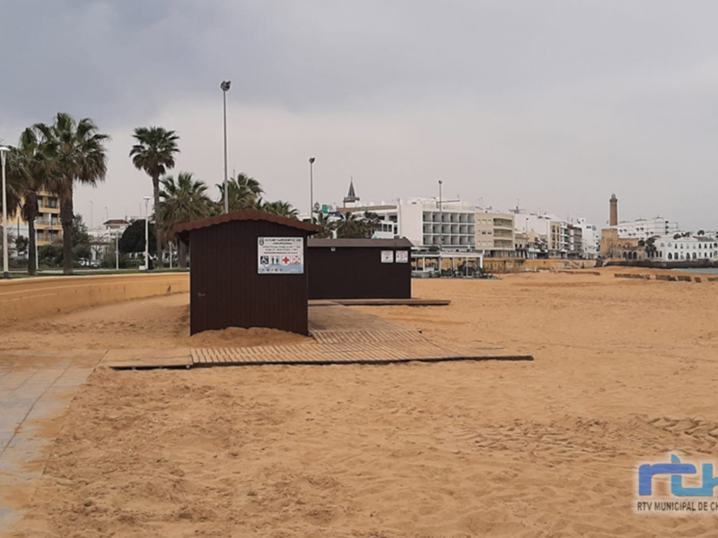 Una pequeña caseta de madera en la arena de una playa, con palmeras y edificios al fondo.