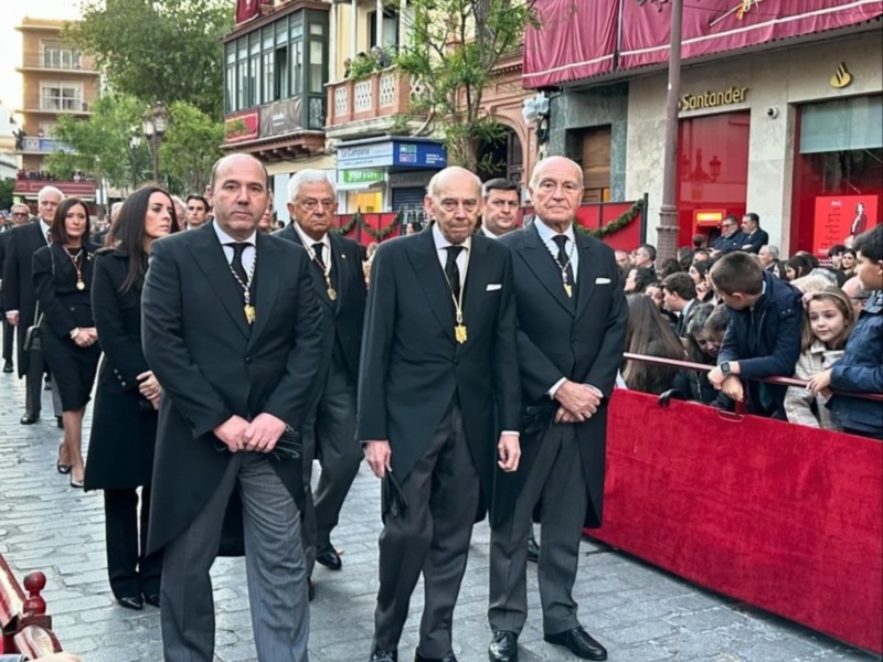 Una fila de hombres en trajes formales caminan por una calle con edificios comerciales a ambos lados. La imagen muestra una celebración o evento formal, con algunos asistentes observando a los hombres en traje.