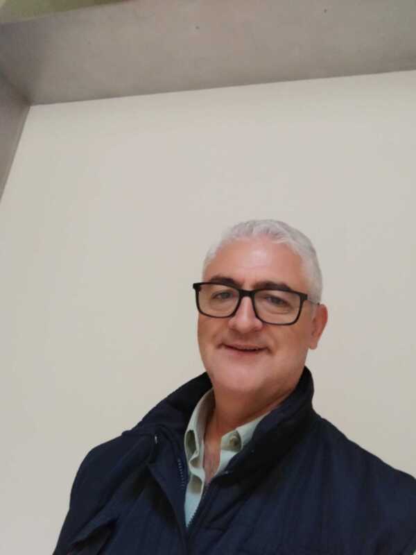 Retrato de una persona con cabello blanco y gafas, vestida con una chaqueta azul y una camisa blanca con detalles en verde y beige.