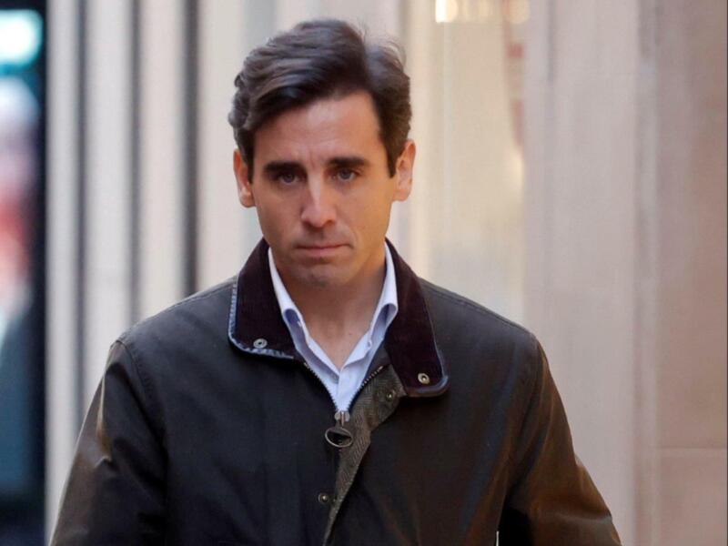 Un hombre con cabello oscuro y barba, vistiendo una chaqueta negra con detalles de color marrón y un cuello azul claro, camina frente a una pared de ladrillos.