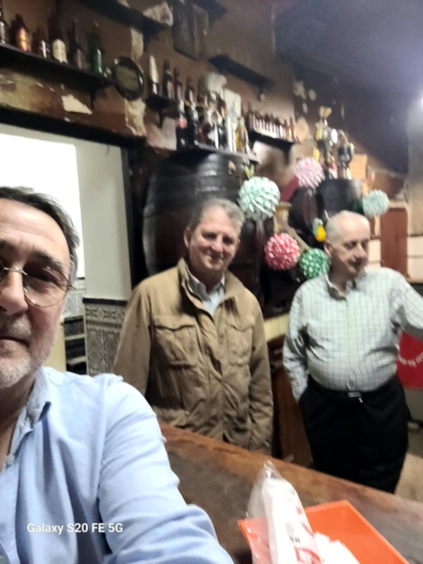Tres hombres sonríen en una barra de bar, con estanterías llenas de botellas y decoraciones festivas en el fondo.