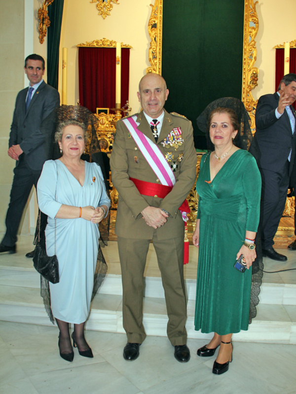 Elegantes individuos posando en un ambiente formal, con vestimentas de gala y adornos de corona. Detalles de la decoración incluyen cortinas rojas y doradas, así como una figura de oro en el fondo.