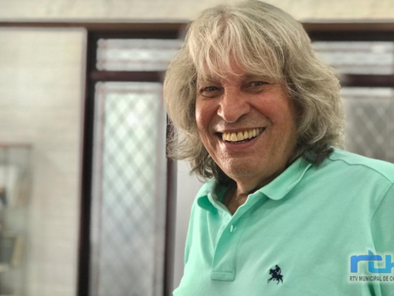 A una persona con cabello largo y gris, sonriendo ampliamente. Se ve vestida con una camiseta verde claro y tiene un logo de caballo en la manga. El fondo incluye una puerta con vidrieras y una estantería de libros.