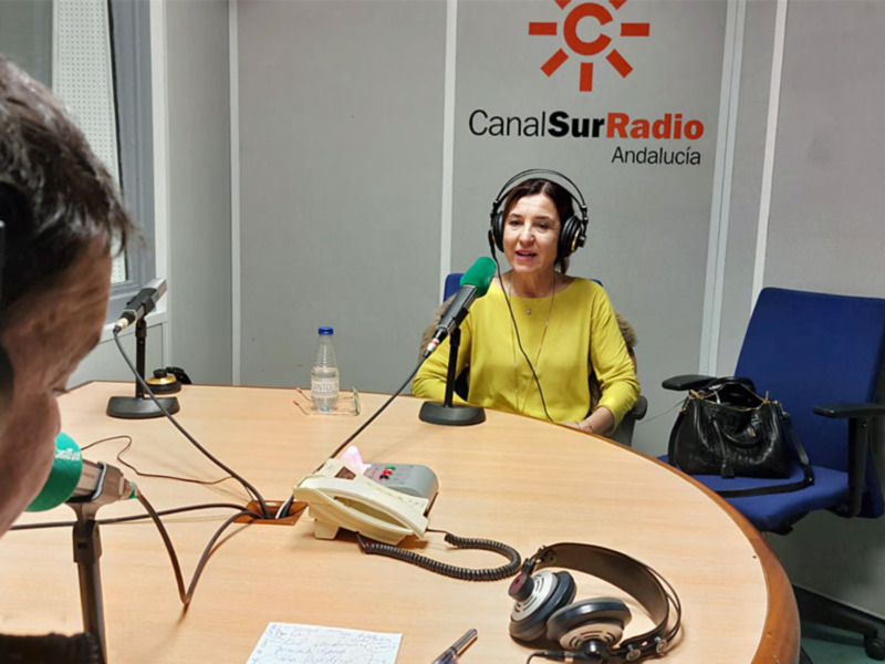 En una sala de radio, dos personas conversan en un estudio profesional. La persona del lado derecho, con auriculares y una botella de agua en la mesa, parece estar en el medio del diálogo. La persona del lado izquierdo, con auriculares y una botella de agua en la mesa, está escuchando activamente. La pared detrás muestra el logotipo de Canal Sur Radio y Andalucía, indicando la ubicación del estudio.