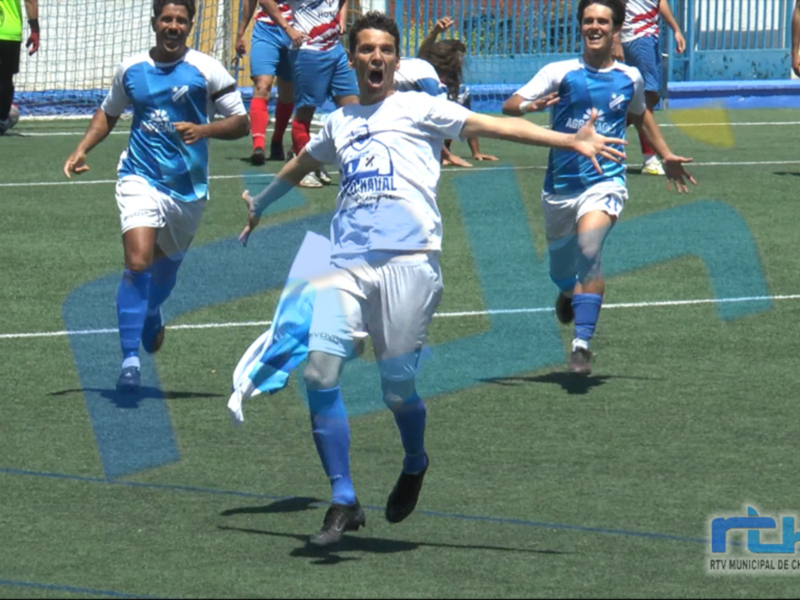 Un jugador de fútbol celebra emocionado en el campo, con otros jugadores al fondo. El estadio es visible con una grada y un marcador en el fondo.
