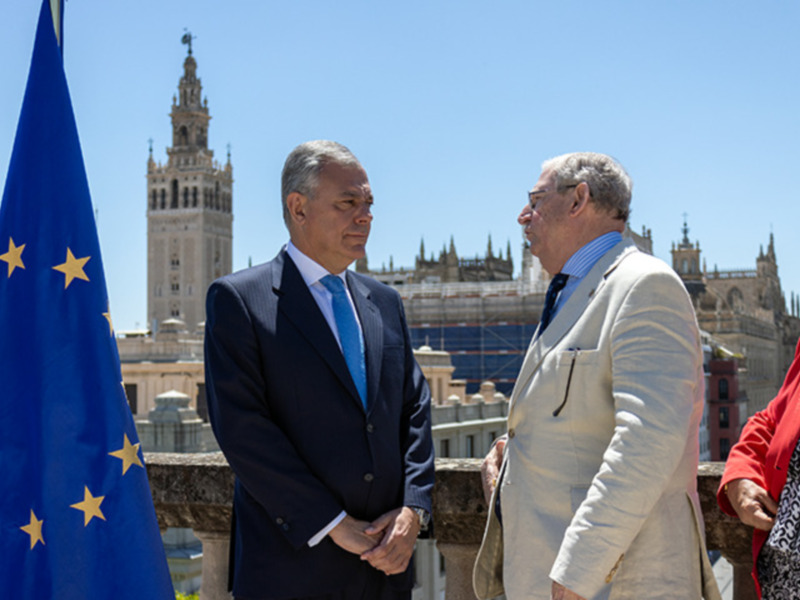 Dos hombres en trajes conversan en un balcón con la bandera de la Unión Europea y la Giralda de Sevilla al fondo.