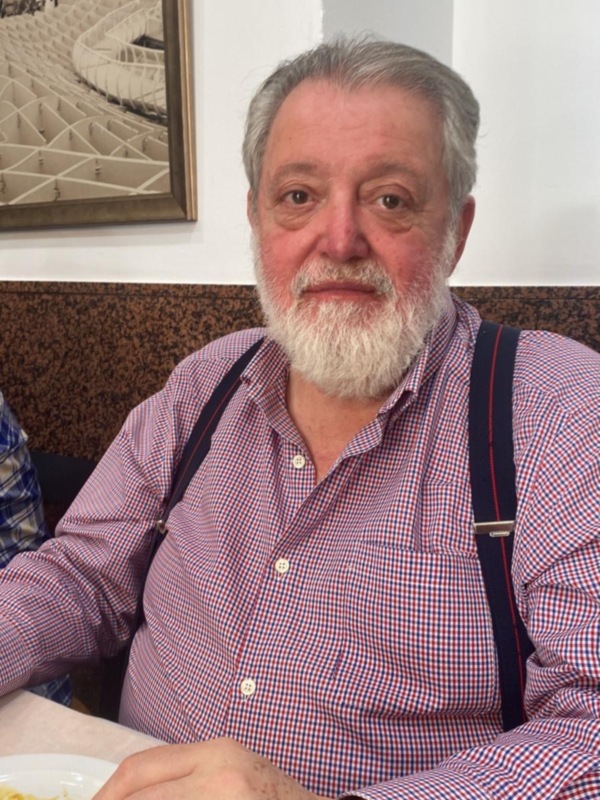 Un hombre con barba blanca y camisa roja está sentado en una mesa, posiblemente en un restaurante. La pared detrás de él tiene un cuadro con una imagen de un estadio deportivo.