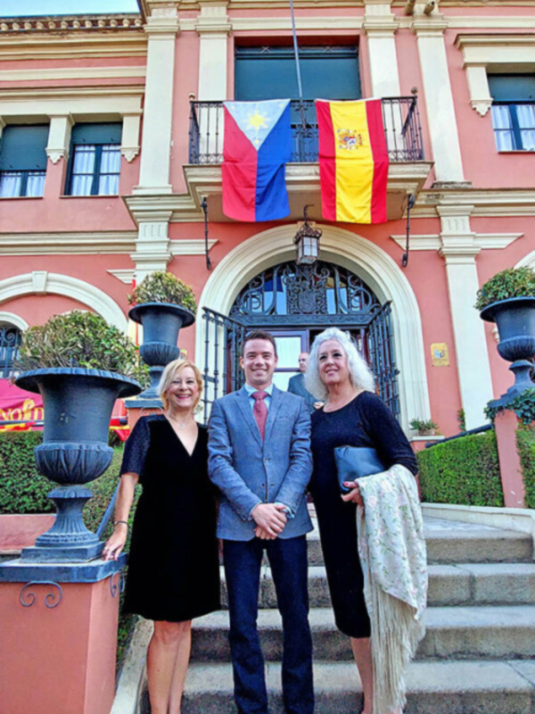 Tres personas posan frente a un edificio con banderas española y de la Comunidad de Madrid, en un ambiente formal. La escena transmite una sensación de orgullo y celebración, posiblemente en el marco de un evento oficial o institucional.
