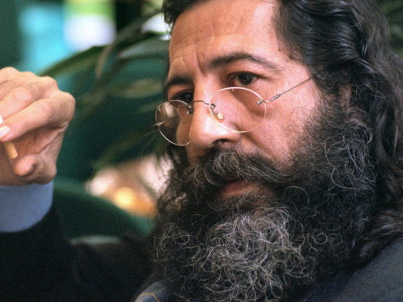 Un hombre con barba y gafas fuma una cigarrilla mientras sostiene la hoja de tabaco.
