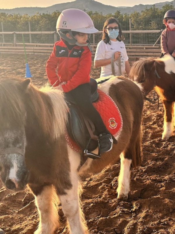 Niños aprendiendo a montar caballos en un campo de arena, con cascos y guantes protectores.