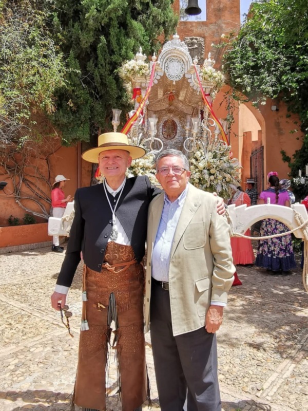 Dos personas posan frente a una carroza decorada con flores y un reloj en el centro, en un entorno con paredes de color naranja y árboles verdes.